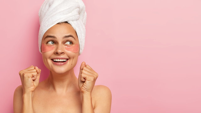 Cropped shot of happy Caucasian woman raises clenched fists, has joyous expression, broad smile, wears hydrogel patches under eyes to reduce wrinkles, isolated over pink wall. Beauty, care concept
