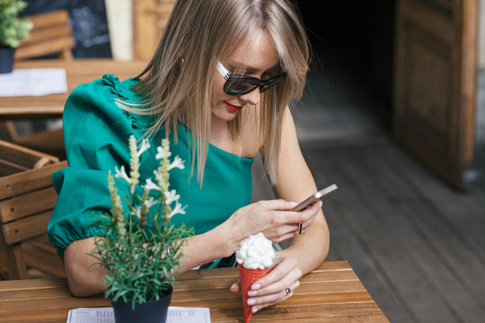 portrait of attractive blonde young woman in sunglasses, green dress and with red lipstick sits at table of street cafe. Woman holding red ice cream cone and looking at cell phone