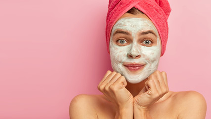Cropped shot of pleased young European female applies facial clay mask, has beauty treatments, cares about complexion and body, stands against rosy studio wall, copy space for your advertisement