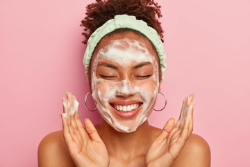 Image of happy ethnic woman raises palms over face, keeps eyes closed, shows white teeth, uses...