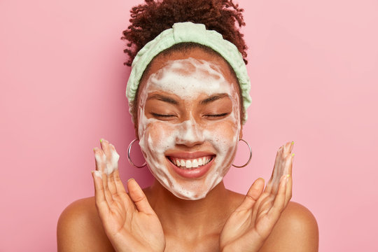 Image of happy ethnic woman raises palms over face, keeps eyes closed, shows white teeth, uses cleansing foam for skin care, gets real pleasure, wears headband and earrings, stands topless alone