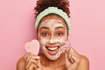 Cropped image of happy African American woman enjoys relaxing time, washes face with soap bubble, feels refreshed and delighted, holds cosmetic sponge for wiping complexion, isolated on pink wall - obrazy, fototapety, plakaty