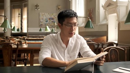 Tracking in shot of young asian male student happily looking in camera reading book in library of university
