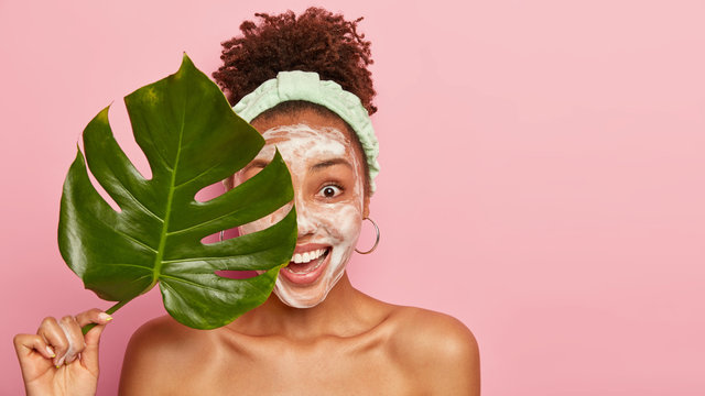 Studio Shot Of Delighted Woman With Curly Hair, Applies Cleansing Foam On Face, Holds Green Plant Leaf, Peels Complexion With Foam, Smiles Broadly, Poses Over Pink Background, Empty Space For Advert