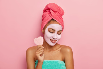 Pleased young dark skinned woman focused down, applies facial clay mask, holds cosmetic sponge for...