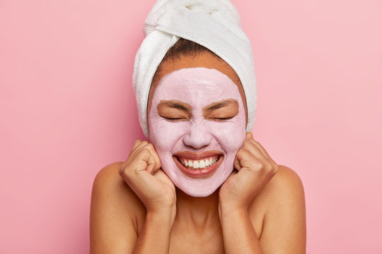 Cropped shot of pleasant looking woman keeps both hands on cheeks, smiles broadly, shows white teeth, wears wrapped towel on head, isolated over pink background, visits cosmetologist in spa centre