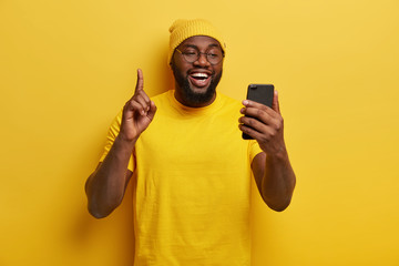 Joyous dark skinned plump man raises index finger, holds mobile phone, enjoys spare time for surfing internet, wears yellow hat and casual t shirt, poses against bright background. Monochrome