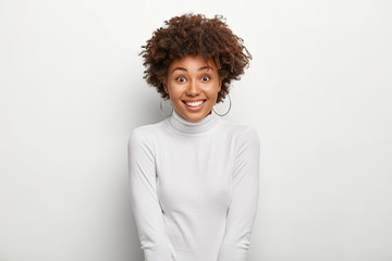 Isolated shot of happy charming woman with Afro hairstyle, wears snow white poloneck jumper, being in good mood after going shopping, poses in studio, expresses happiness. Positive emotions concept
