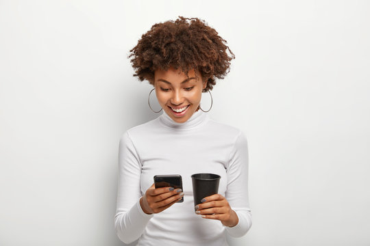 Photo of happy African American teenage girl surfes internet on mobile phone, connected to free wifi, drinks takeout coffee, wears casual turtleneck jumper, creats own blog, being always in touch