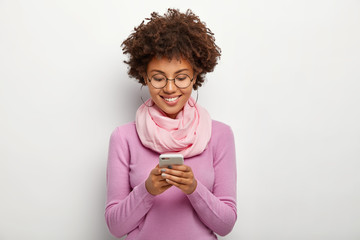 Pleased dark skinned woman focused in smartphone device, has cheerful expression, checks...