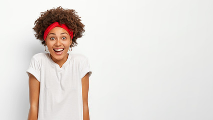 Portrait of happy dark skinned woman with curly Afro hairstyle, dressed in casual wear, expresses positive emotions, laughs at something funny, isolated over white background. Wow, its great!