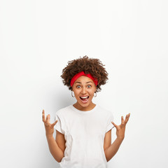 Excited young dark skinned woman with curly crisp hair, gestures actively, laughs out, gets pleasant emotions from seeing something cool and pleasant, dressed in casual wear, isolated on white wall