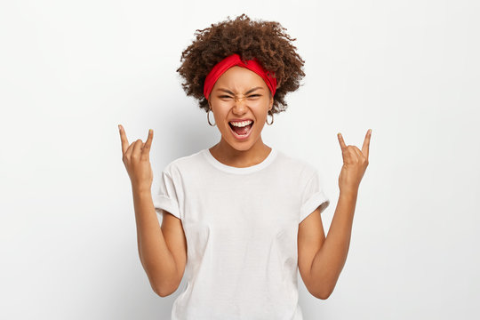 Photo of energetic young African American woman makes rock n roll gesture, enjoys cool music, smirks face, dressed in casual wear, poses against white background. People, body language concept
