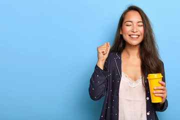 Joyous happy Asian woman with long dark hair, enjoys morning coffee, clenches fist, dressed in nightwear, smiles broadly, poses against blue background, rejoices this day started. Awakening concept