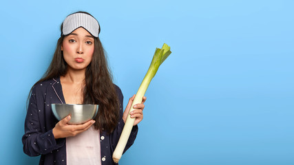Studio shot of upset housewife wakes up early to prepare breakfast, holds fresh vegetable and bowl,...