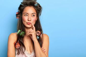 Pensive beautiful woman with Asian appearance, wears hair curlers, makes hairstyle before date,...