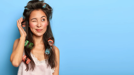 Studio shot of thoughtful Korean woman prepares for party, wears hair curlers for making hairstyle,...