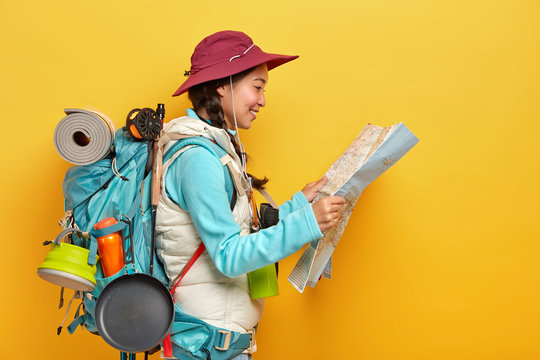 Sideways Shot Of Asian Female Tourist Studies Map, Finds New Destination To Explore, Travels Alone, Wears Cap And Active Wear, Carries Big Rucksack With Necessary Things For Travel, Isolated On Yellow