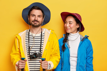 Two mixedrace woman and man have active rest, trek together, explore new destination, use special trekking sticks, wear hats, use retro camera for making photos during travel, have happy moods