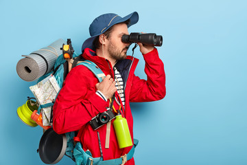 Photo of male explorer dressed in casual wear, keeps binoculars near eyes, wears hat and jacket,...