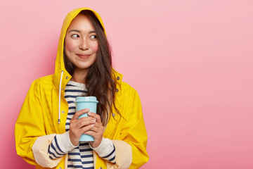 Portrait of cute young woman wears raincoat, warms with hot beverage, looks happily aside, has dimples on rouge cheeks, enjoys rainy weather in comfortable streetwear, isolated on pink wall.