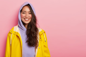 Young girl with dark hair, wears purple hoody, dressed in yellow raincoat, smiles pleasantly, poses against pink background, copy space area aside for your promotional content. Rainy weather concept