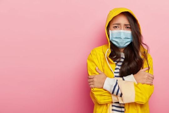 Upset Asian woman trembles from cold, has virus transmitted through airbone droplets, wears protective medical mask, yellow raincoat with hood, stands against pink wall, has contagious disease
