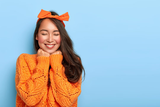 Portrait of cheerful mixed race woman has shy satisfied expression, smiles broadly, shows white teeth, wears orange bow headband and knitted sweater, poses against blue background. Ethnicity, emotions