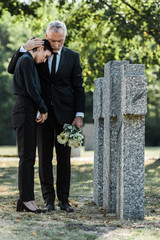 senior man holding flowers and hugging upset woman near tombstones