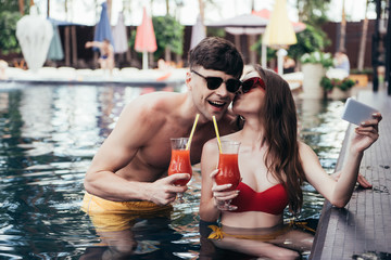 happy young woman taking selfie while kissing boyfriend in swimming pool