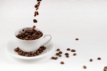 A cup with coffee beans in the air flying in flight like a spray. On background of a white wooden table, coffee shop advertising concept, copy space.