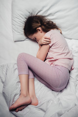 Naklejka premium top view of scared child lying in bed and hugging herself with arms
