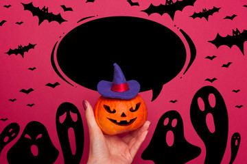 Happy halloween Pumpkin hold in a female hand in a blue witch hat with a white hand lettering in a black speech bubble. Black silhouettes of ghosts and bats.