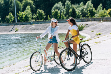 Fototapeta na wymiar happy blonde and brunette girls with bikes looking at each other near river in summer