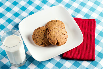 Healthy grain cookies served on white plate with a glass of milk on a table with light blue checkered tablecloth