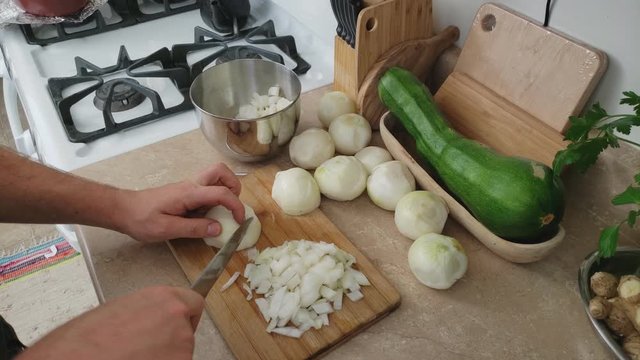 Home cooking - High angle view of spiting and mincing more onion on small bamboo cutting board using chef knife while more onion and large zucchini on the counter top.