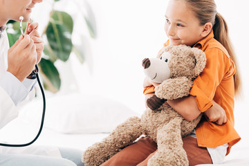 partial view of pediatrist in white coat and kid with teddy bear in clinic