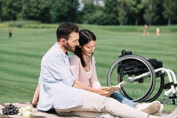 Naklejka premium happy reading book with disabled girlfriend while sitting on blanket in park