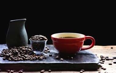 Selective focus at red coffee cup with milk jug and coffee beans on black slate and wooden table in dark background