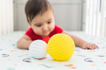 Baby playing with colorful toy rubber balls in the crib in a bright room. Child wearing a red bodysuit, laying on playful dinosaur crib sheet.
