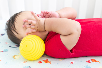 Baby playing with colorful toy rubber balls in the crib in a bright room. Child wearing a red bodysuit, laying on playful dinosaur crib sheet.