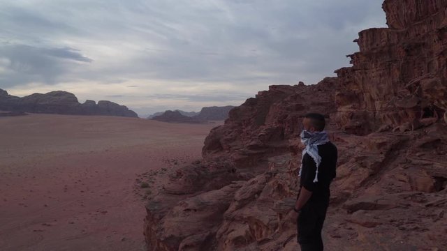 man walking towards the edge of a cliff in the desert of wadi rum and covering his face with the arabic scarf