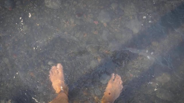 POV, Medium shot, Waves crashes onto the feet of a young man and ebbs away as he stands on a stony beach. 