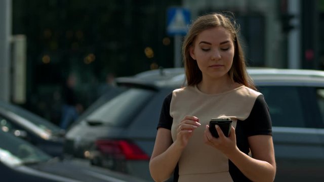 Teenage girl dressed up texting on her phone in city environment, cars driving behind her. She is oblivious of the traffic. Slow motion shot.