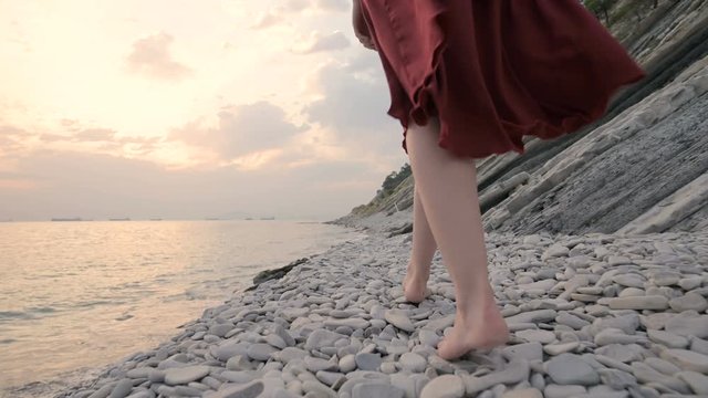 Close-up of the legs of a young girl with a red dress from behind walking on a rocky beach the sea coast at sunset. A dress fluttering in the wind in the wind.