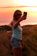 at orange sunset by the sea on a hill there is a girl with long hair in shorts and a T-shirt with a phone, takes a selfie