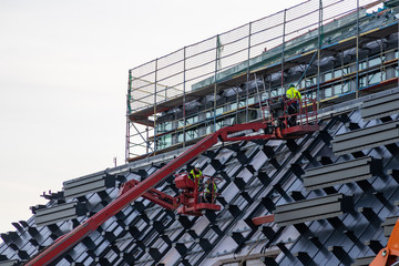 Baustelle mit Bauarbeitern auf roten Hebebühnen und roten Kränen begutachten die Fassade, die Glasfassade und das Gerüst der Architektur anhand der Baupläne