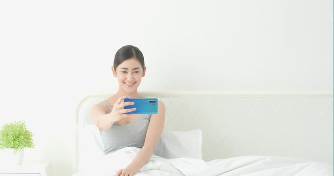 Asian woman posting and take photo for new picture of yourself with  smartphone at bed room. Concept of technology, communication, webcam, uploaded and selfie.