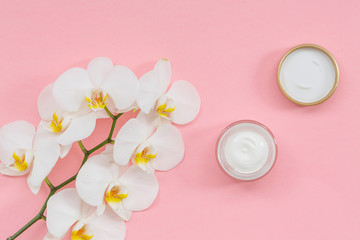 Opened plastic container with cream and White Phalaenopsis orchid flowers on pink background. Flat lay. Natural beauty product for skin care. Organic cosmetics concept.