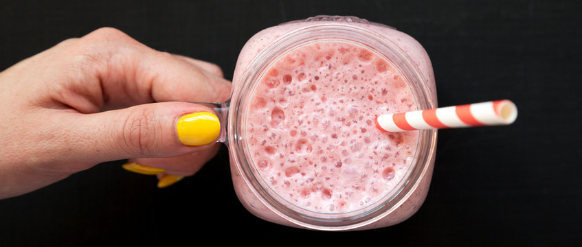 Female hand holding a glass jar filled with strawberry banana smoothie over black surface, overhead view. Top view, from above, flat lay. Closeup.